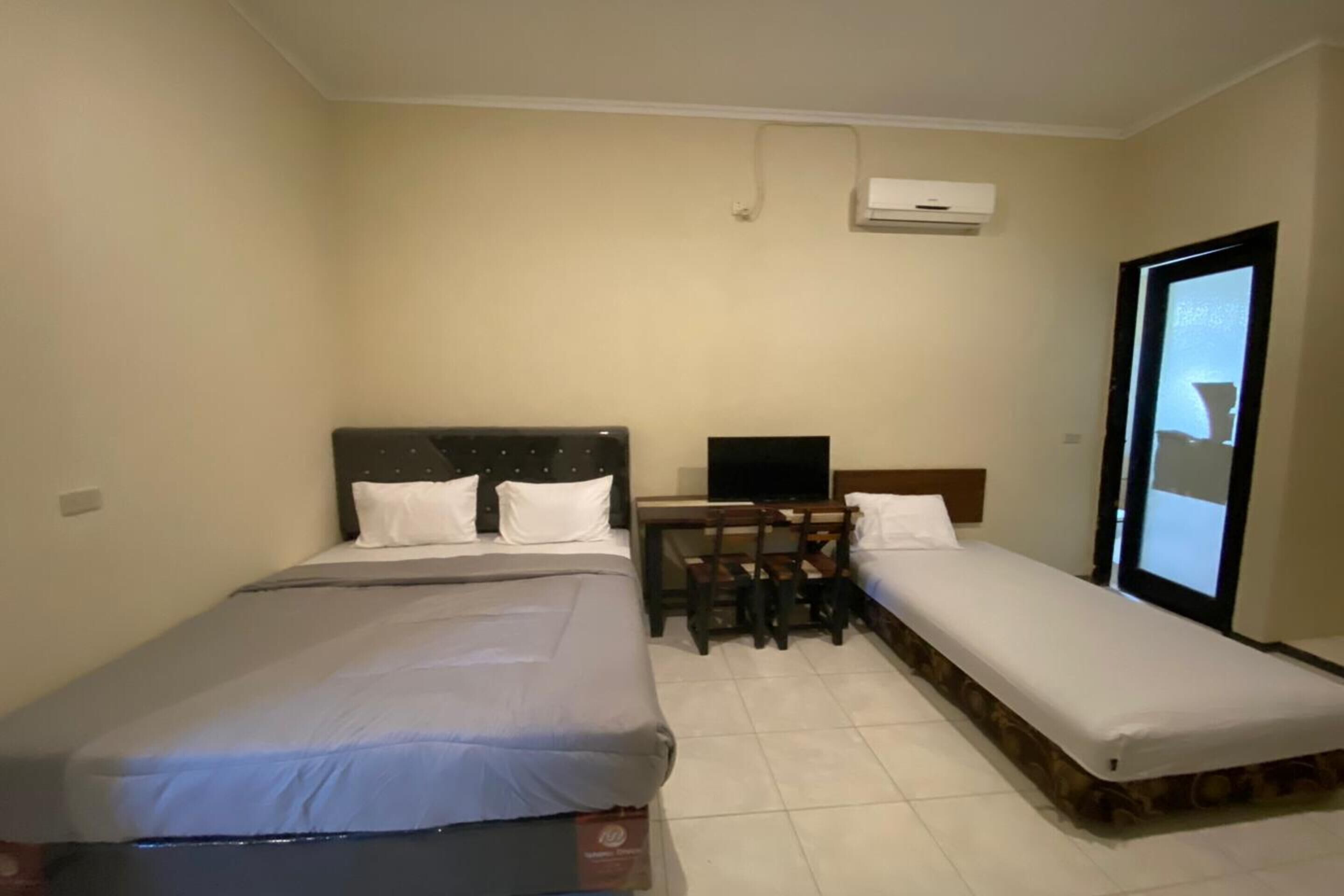 A hotel room with a large bed, a smaller bed, a desk with a laptop, and an air conditioning unit on the wall.