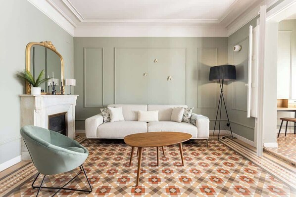 A living room with a white sofa, a green armchair, a wooden coffee table, a fireplace, and a patterned floor.