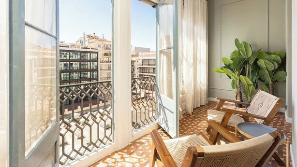A balcony with a view of buildings, a patterned floor, and two wooden chairs with cushions. - Spacious Stylish 4-BR Modernist Apartment (Barcelona)