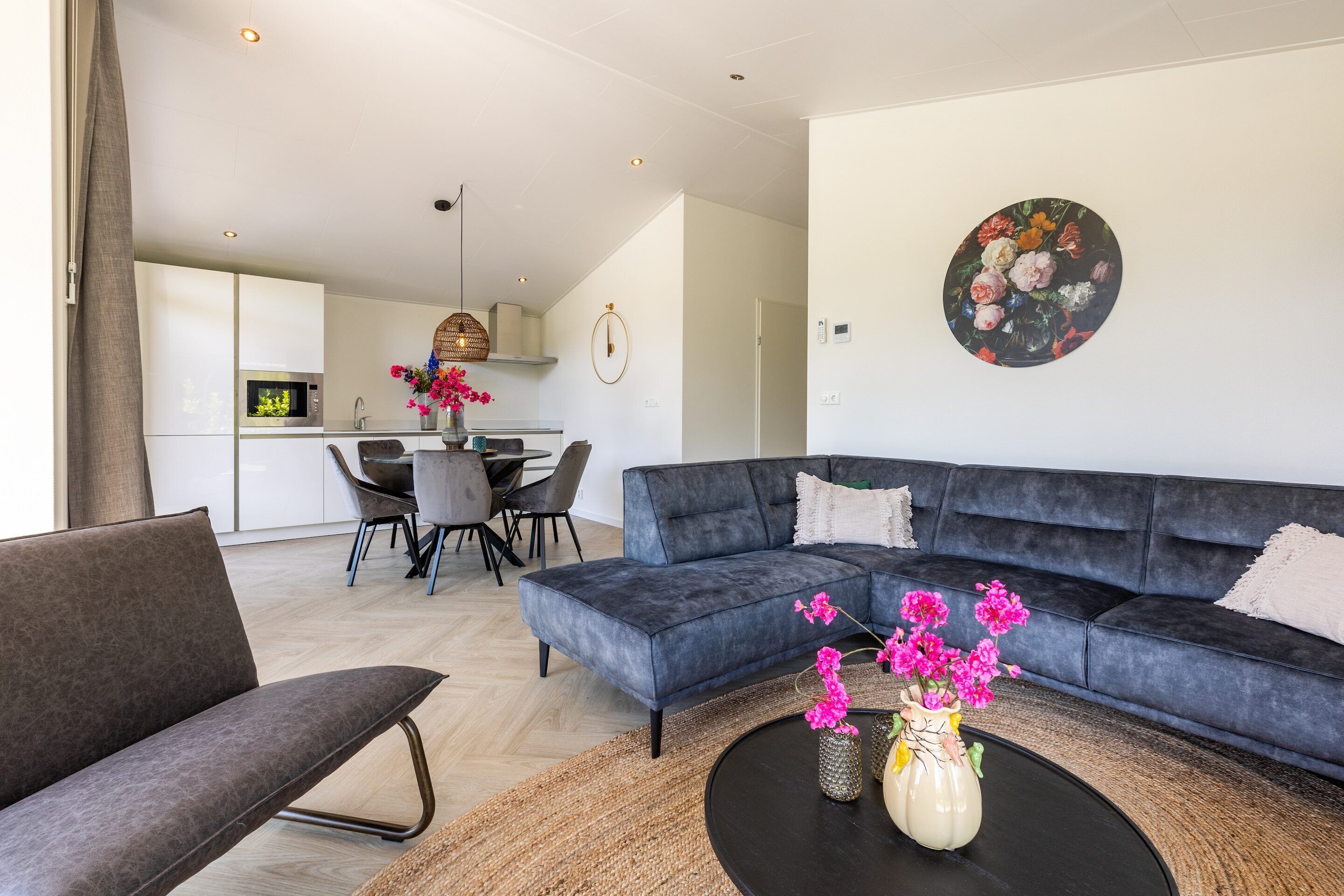 Een moderne woonkamer met een donkergrijze bank, een ronde salontafel met een bloevas, en een eethoek op de achtergrond.