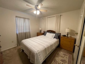 A bedroom with a bed, a ceiling fan, a nightstand, a lamp, and a window with curtains.