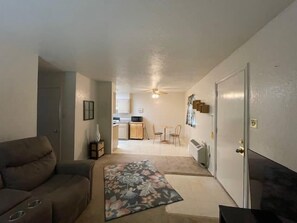 A living room with a sofa, a coffee table, and a door leading to a kitchen area with a dining table and chairs.
