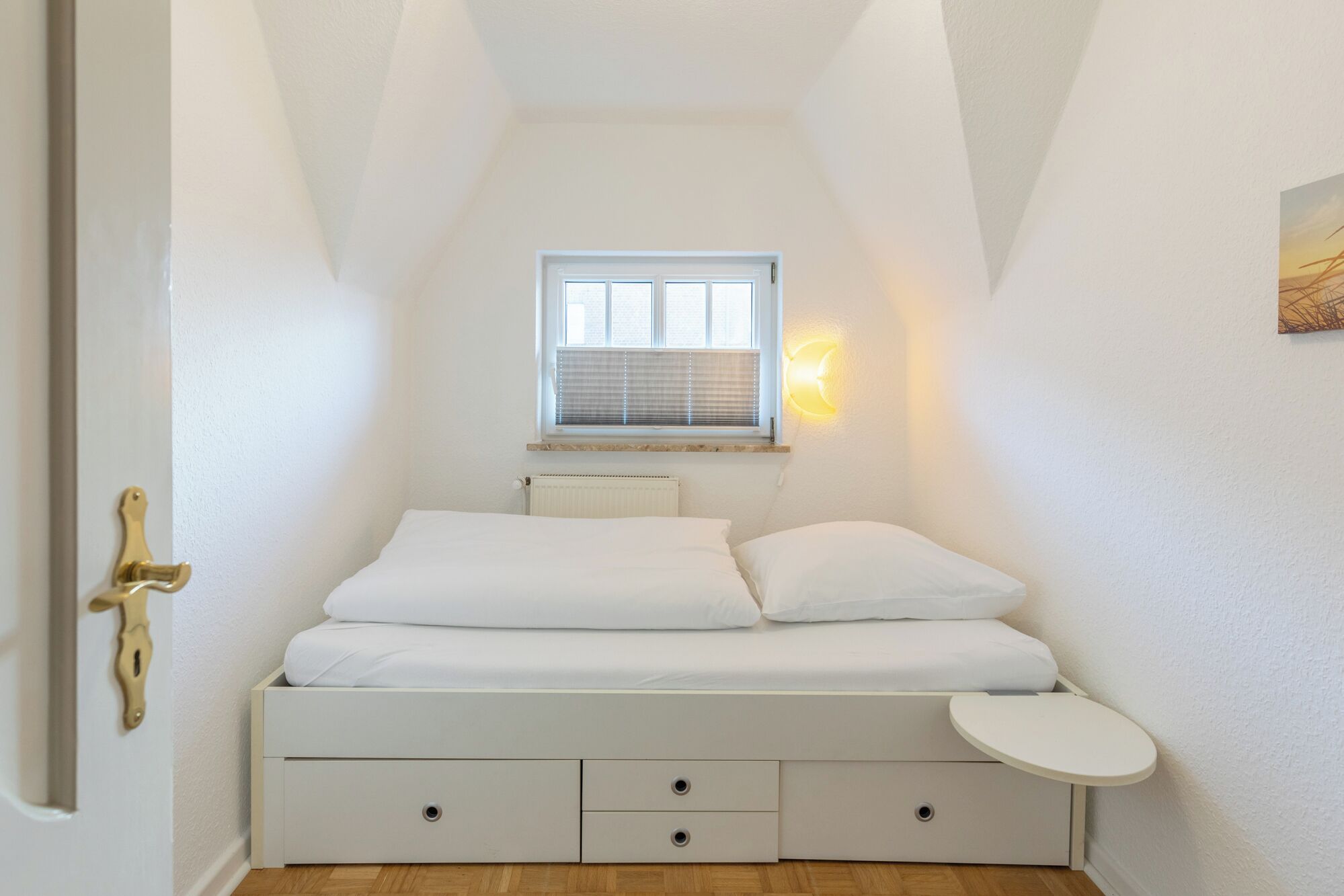 Une petite chambre blanche avec un lit simple, une fenêtre, une table de chevet et un radiateur.