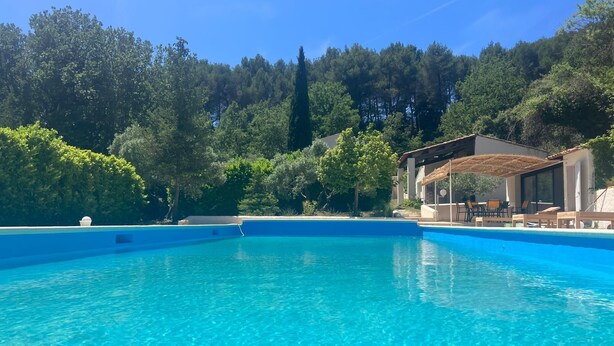 Una piscina azul cristalina con una zona de estar cubierta y un edificio al fondo.