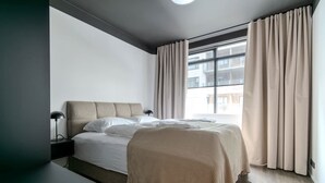 Habitación de hotel moderna con una cama grande, una mesita de noche, una lámpara y vistas a la ciudad desde la ventana.