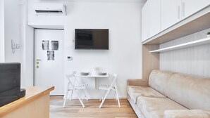 A compact living space with a kitchenette, a flat-screen TV mounted on the wall, a dining table with chairs, and a beige sofa.