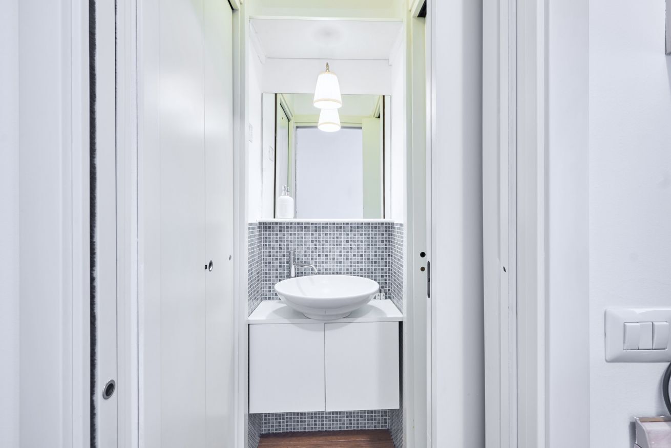 A narrow hallway with a white sink and mirror on a tiled wall, a door on the left, and a light switch on the right.