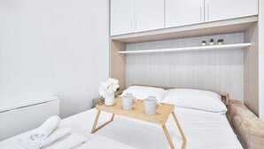 A neatly made bed with a wooden tray holding two mugs and a vase of flowers, a white towel, and a small plant on a shelf above the bed.