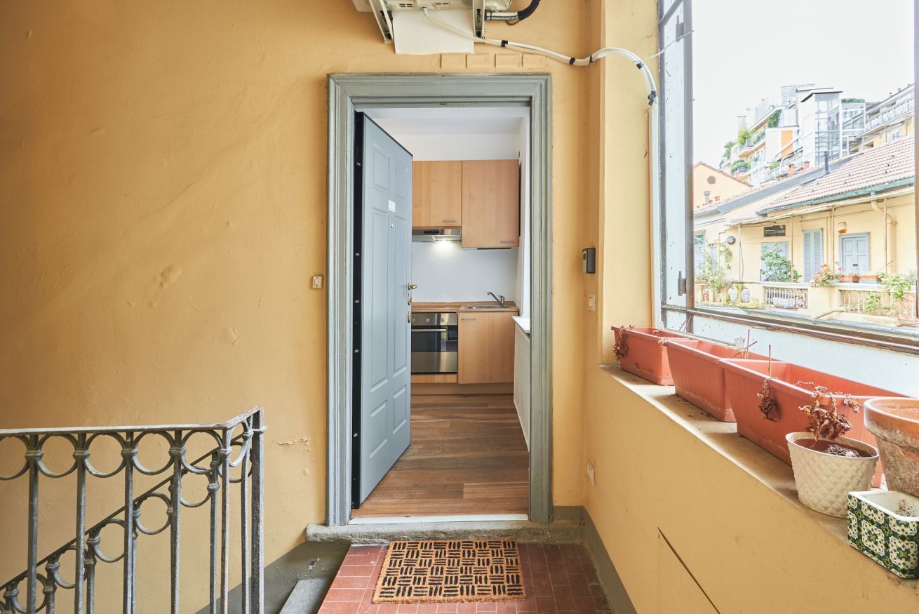 A narrow hallway leading to a kitchen with wooden cabinets and a window overlooking a balcony with potted plants.