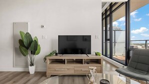 Uma sala de estar moderna com uma TV de tela plana sobre um console de madeira, uma mesa de centro de vidro, uma planta em vaso e vista da cidade através de grandes janelas.