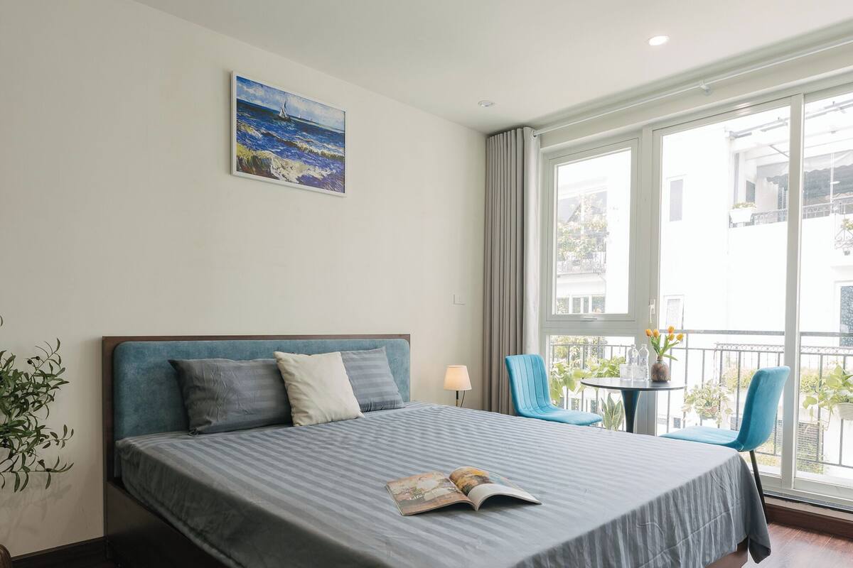 A modern hotel room with a large bed, a small table with chairs, a balcony view, and a potted plant.