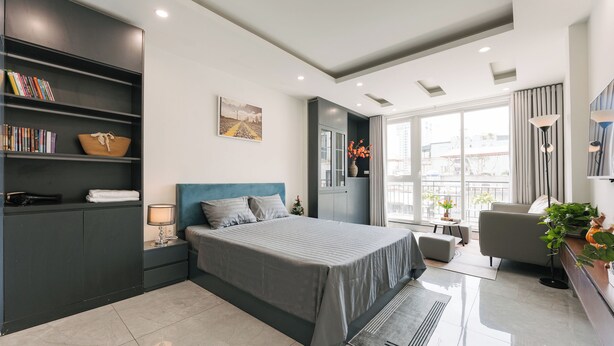 Un dormitorio moderno con una cama grande, estanterías empotradas, una mesita de noche, una lámpara y vistas a la ciudad a través de amplios ventanales.