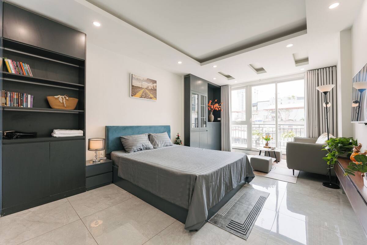 A modern bedroom with a large bed, built-in shelving, a bedside table, a lamp, and a view of the city through large windows.