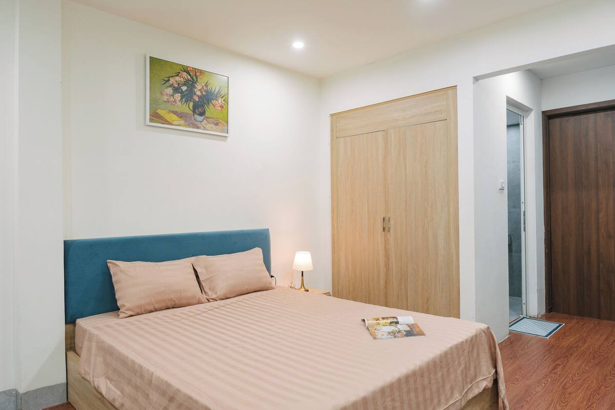 A bedroom with a bed, a nightstand with a lamp, a wooden wardrobe, and a door leading to another room.