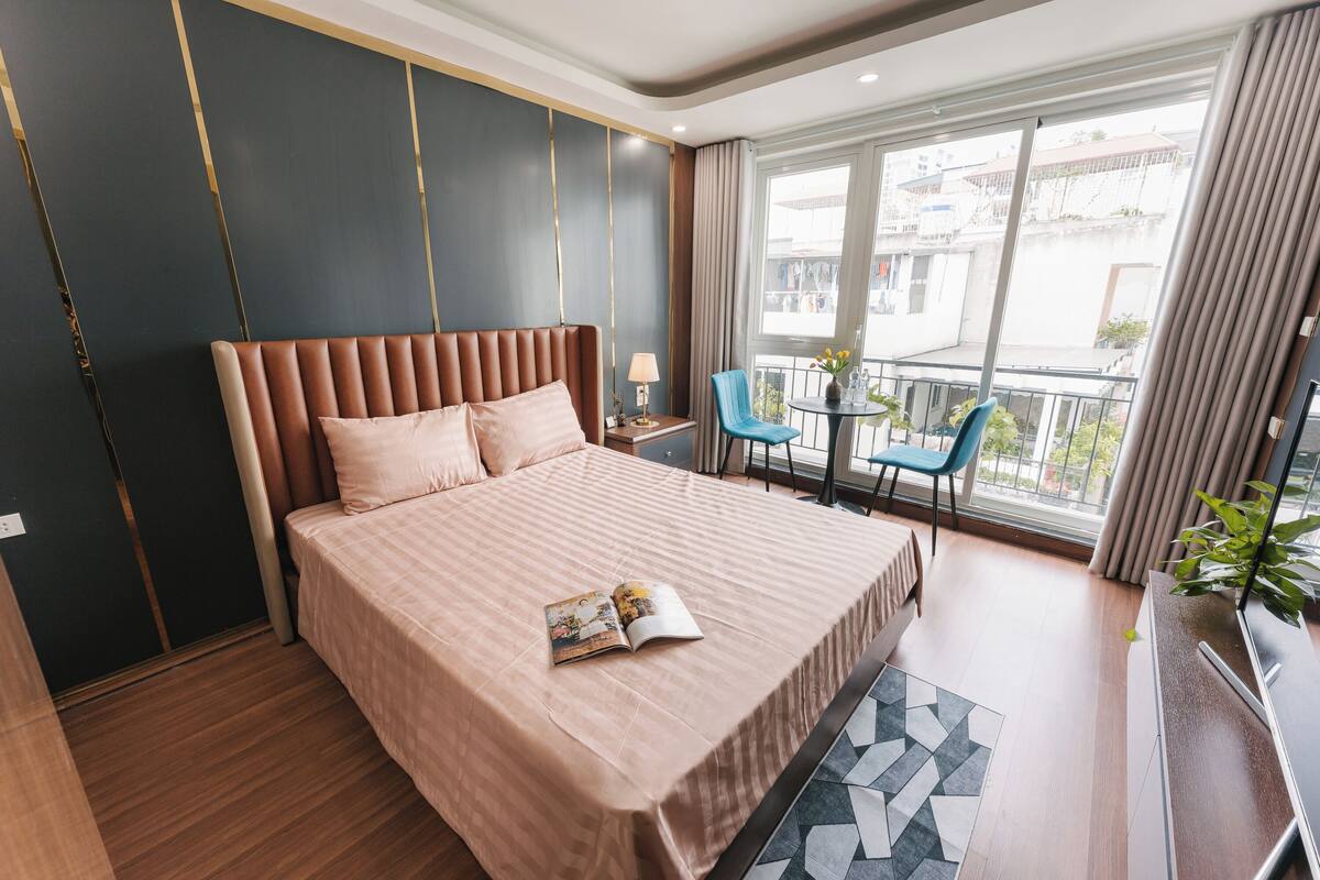 A modern hotel room with a large bed, a small table with a lamp, a chair, and a balcony with a view of the city.