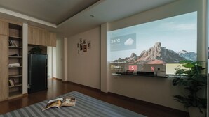 Un salón moderno con un televisor grande mostrando un paisaje montañoso, una estantería, un refrigerador y una planta en maceta.