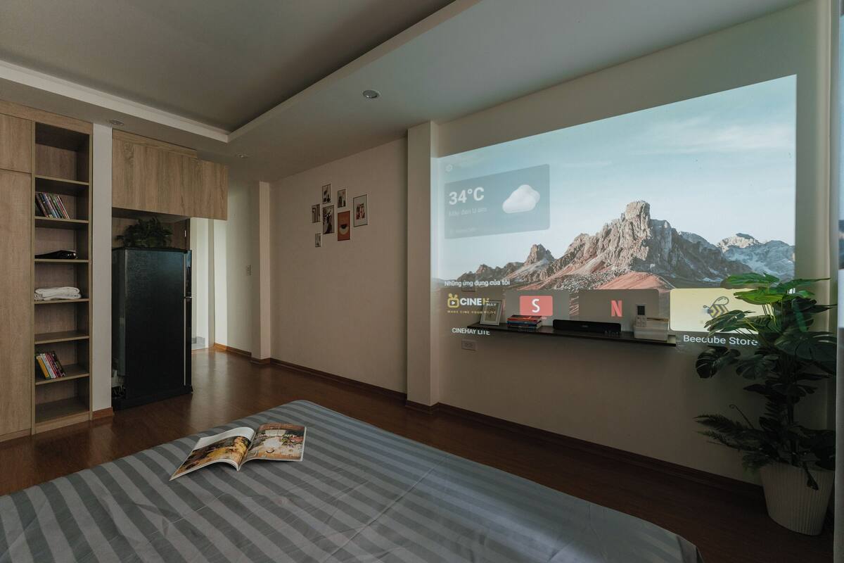 A modern living room with a large TV displaying a mountain landscape, a bookshelf, a refrigerator, and a potted plant.