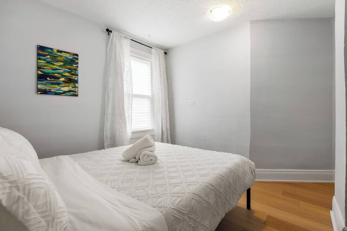 A bedroom with a bed, a window with sheer curtains, a painting on the wall, and a wooden floor.