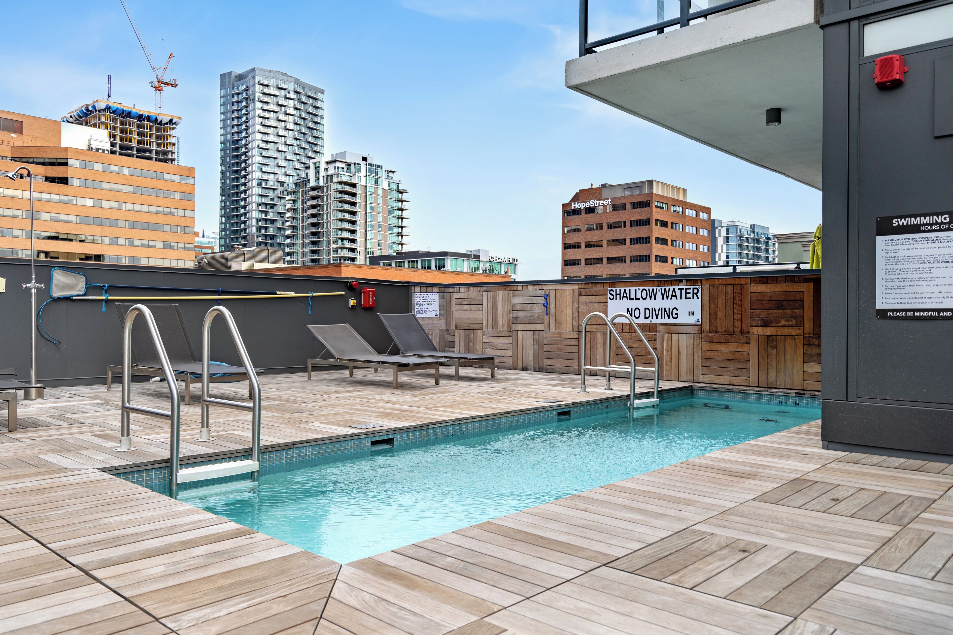 Une piscine sur le toit offrant une vue sur la ville, un panneau indiquant la faible profondeur de l’eau et des chaises longues.