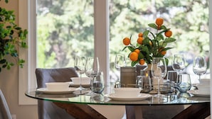 Ett matbord dukat med genomskinliga glas och tallrikar, en vas med orange blommor och en utsikt över träd genom stora fönster.