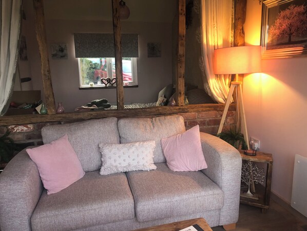 Un salon chaleureux avec un canapé gris, des coussins roses, un plafond avec une poutre en bois, une fenêtre donnant sur un beau paysage et une lampe sur une table d’appoint.