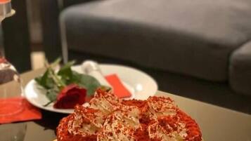 Un pastel redondo con glaseado rojo sobre una mesa en una habitación con un sofá gris y una mesita con un mantel individual rojo.