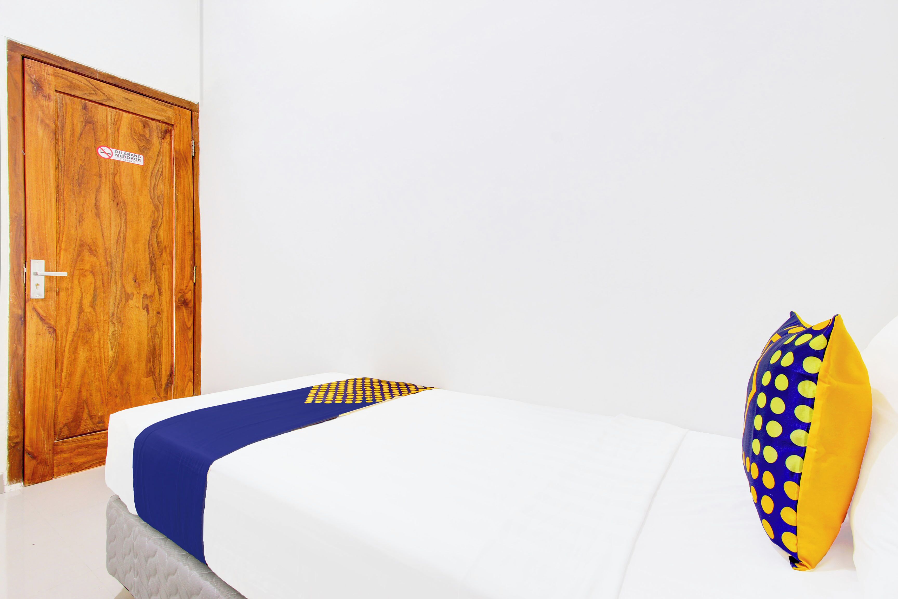Una habitación con puerta de madera, cama con ropa de cama blanca y almohada azul y amarilla, y una manta a cuadros.