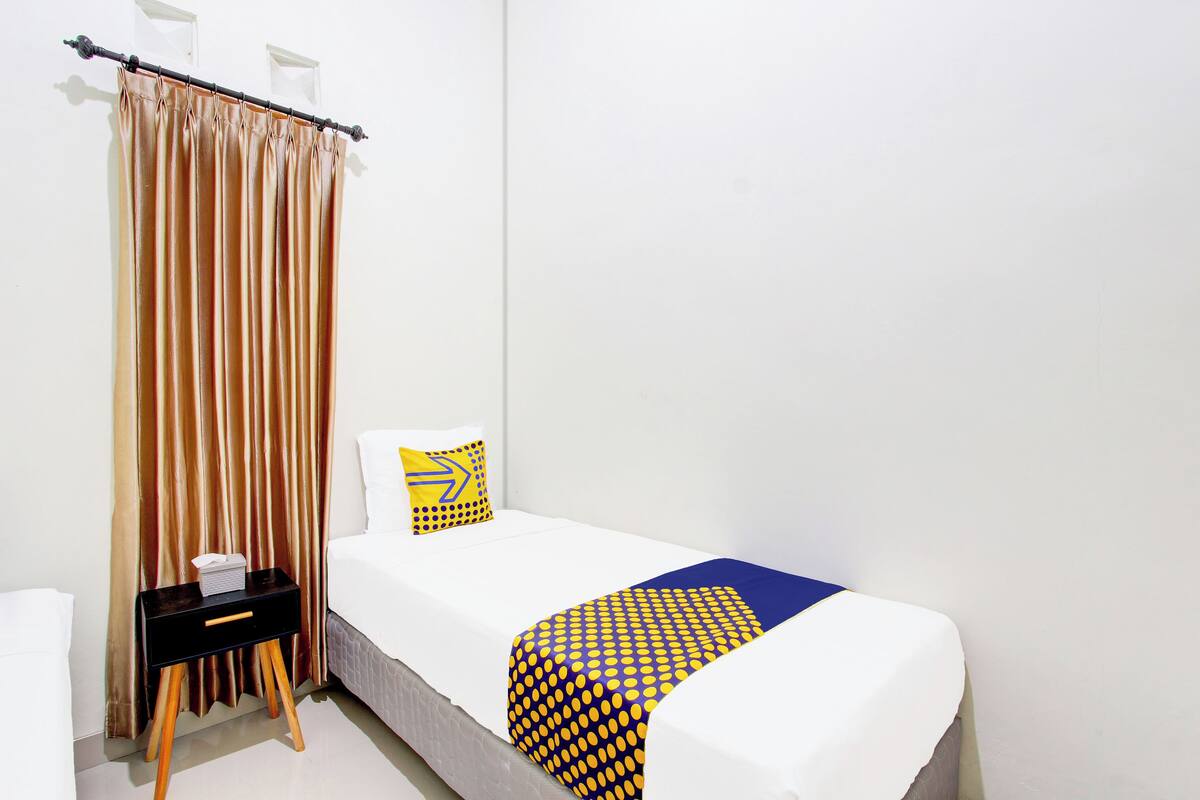 Una cama pequeña y bien hecha con colchón blanco y una manta de colores azul y amarillo. Hay una mesita de noche negra con una caja de pañuelos blancos y un almohadón amarillo con diseño azul. Una cortina con rayas verticales está parcialmente recogida.