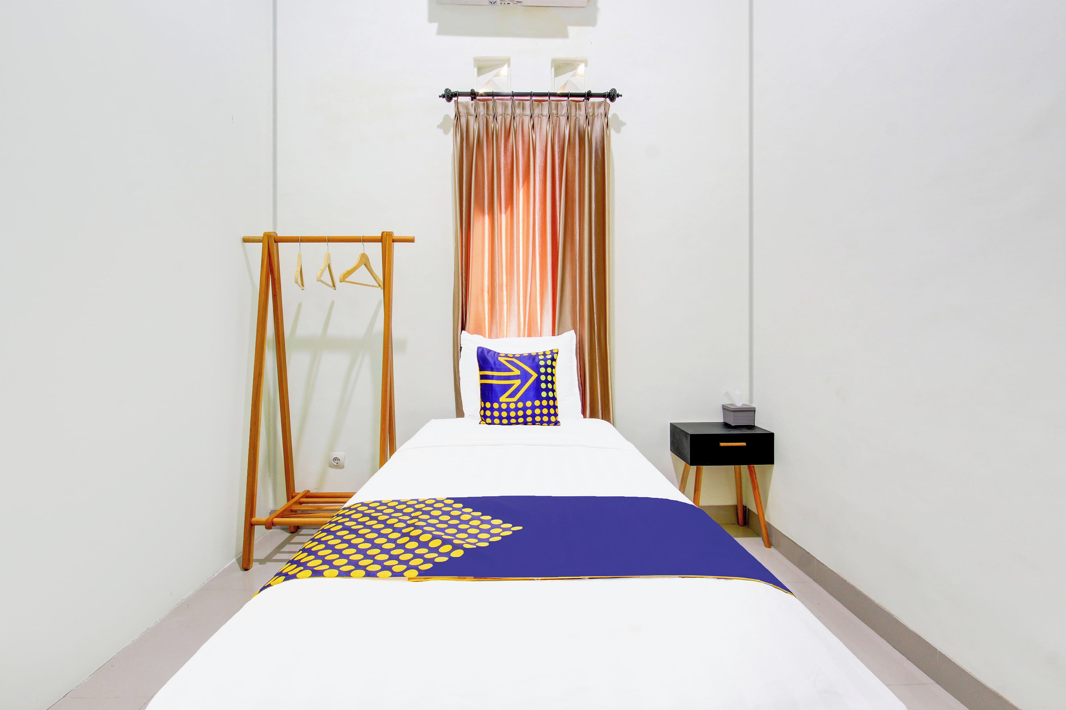 A neatly made bed with a blue and yellow patterned bedspread, a wooden drying rack, a black bedside table, and a window with curtains.
