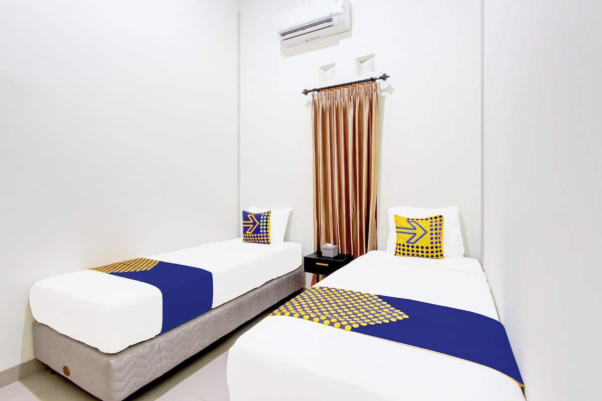 Habitación de hotel con dos camas individuales, cada una con colchón blanco, cubrecamas estampadas en azul y amarillo, y almohadas a juego. Hay una mesita de noche negra pequeña con una lámpara y un equipo de aire acondicionado blanco sobre las camas.
