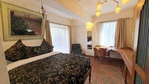 A room with a bed, a desk, and a chair. There is a window with curtains, a chandelier, and a painting on the wall. - Woodmere Grand Hotel (Nairobi)