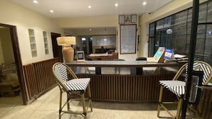 A reception area with a counter, two chairs, a lamp, and a menu board. - Woodmere Grand Hotel (Nairobi)