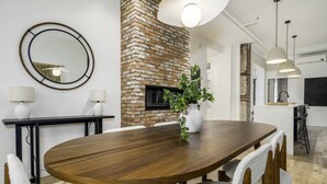 Un comedor moderno con mesa de madera, sillas, pared de ladrillo decorativa, televisor, lámparas colgantes y una planta.
