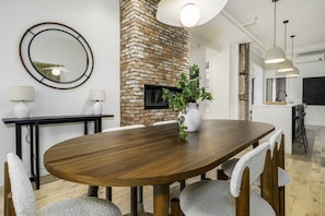 A modern dining area with a wooden table, chairs, a brick accent wall, a TV, pendant lights, and a plant.
