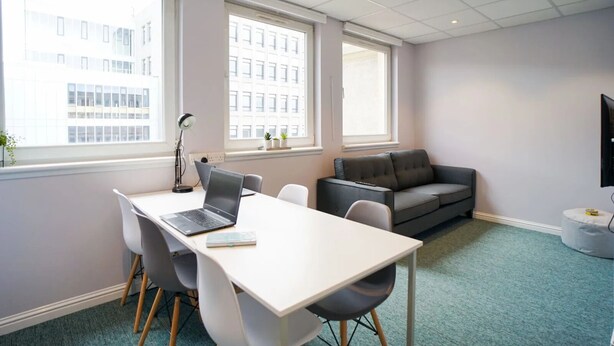 Ein moderner Arbeitsraum mit weißem Schreibtisch, Laptop und Stühlen, einer grauen Couch und einem Fernseher an der Wand.