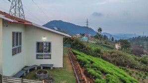 A house with a sloped roof, a small garden with a fire pit, and a view of a hilly landscape with power lines and a distant building. - The springs Nilgiris (Kotagiri)