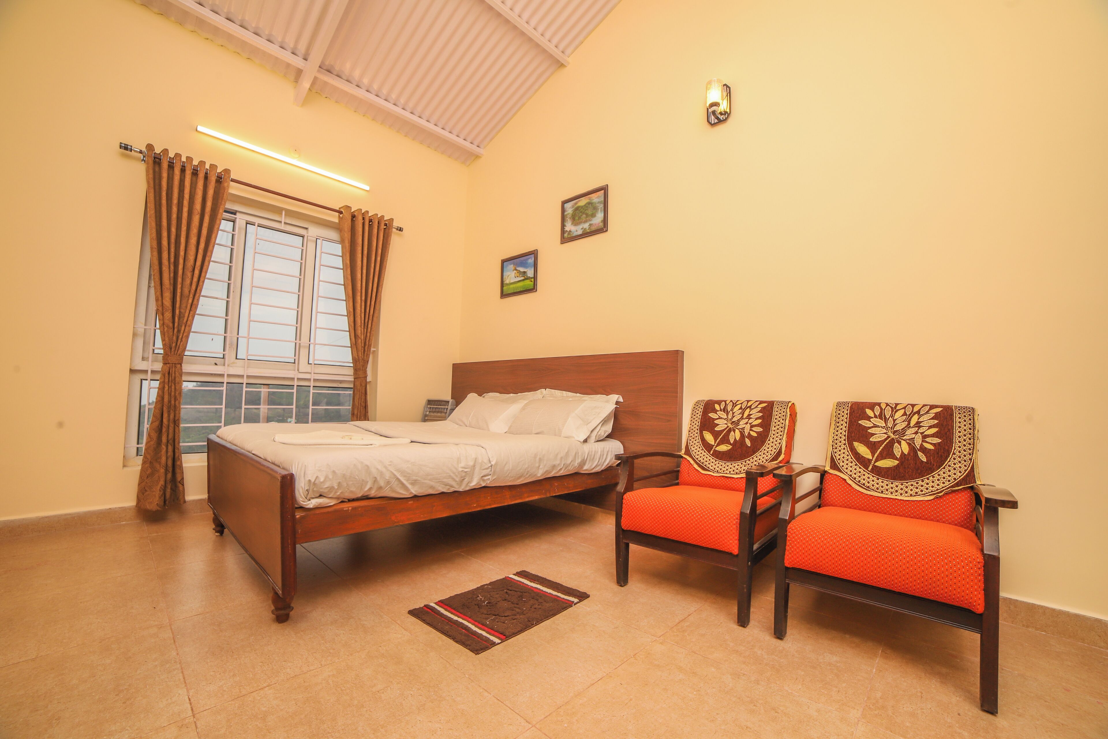 A bedroom with a bed, two chairs, a window with curtains, and a wall-mounted light fixture.