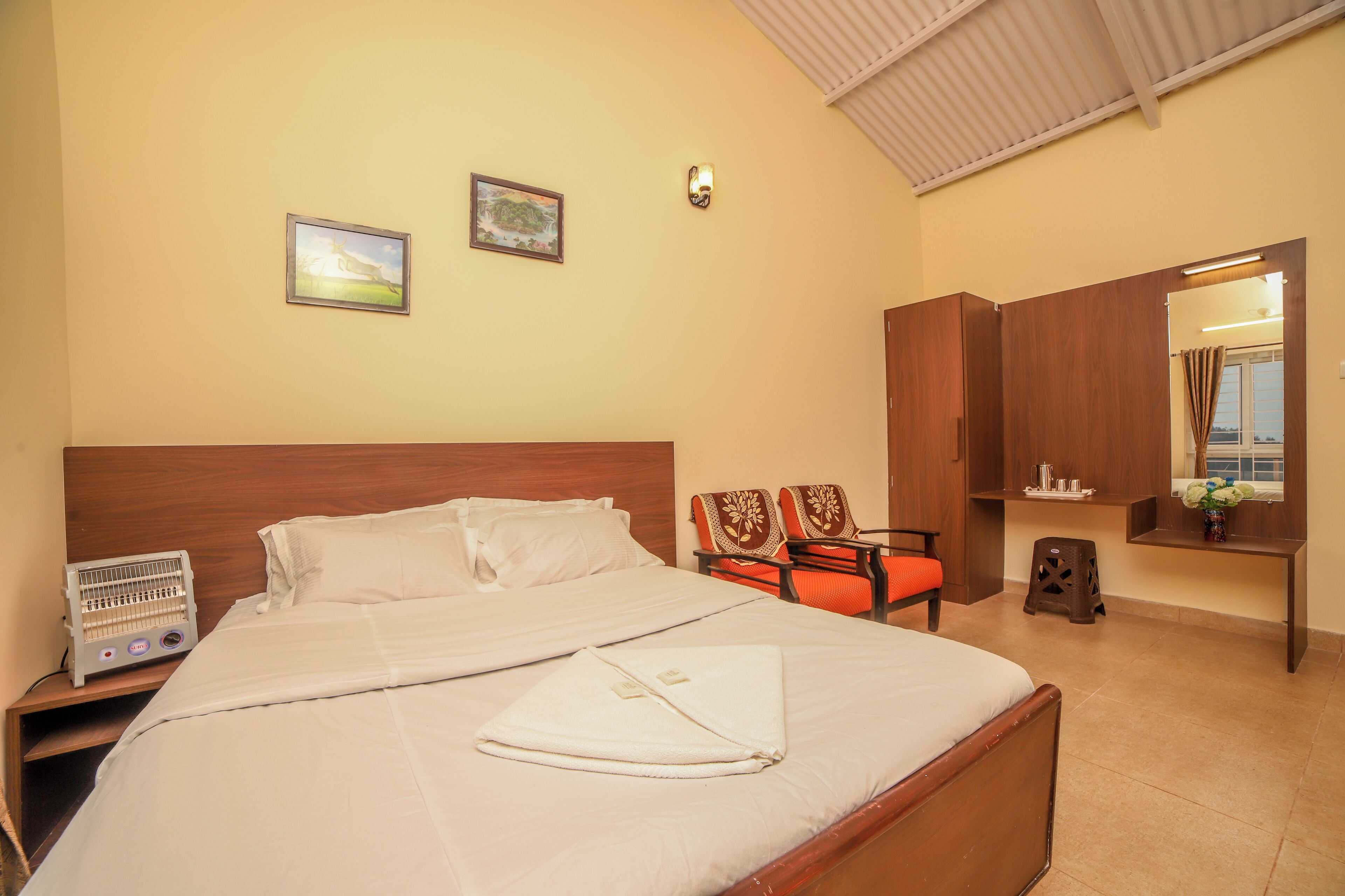 A neatly made bed with a white comforter and pillows, a bedside table with a fan, a wooden headboard, a mirror, and a small stool.