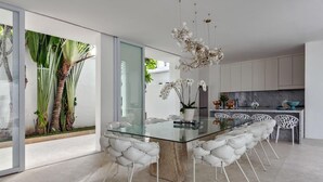 Un comedor moderno con una mesa de vidrio y sillas blancas acolchadas, una cocina con armarios blancos y vistas a plantas tropicales.