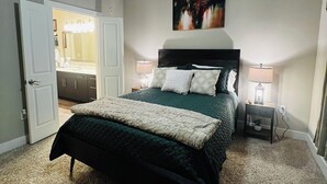 Ein Schlafzimmer mit Bett, Deckenventilator, Nachttisch mit Lampe, einem Bild an der Wand und einer offenen Tür zu einem Badezimmer.