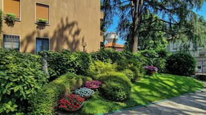 A well-maintained garden with various plants and flowers, a statue, and a building with multiple windows and balconies. - Rovani 14 (Monza)