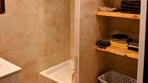 Une salle de bain carrelée de carreaux beiges, avec une baignoire, une étagère où sont pliées des serviettes et un panier à linge.