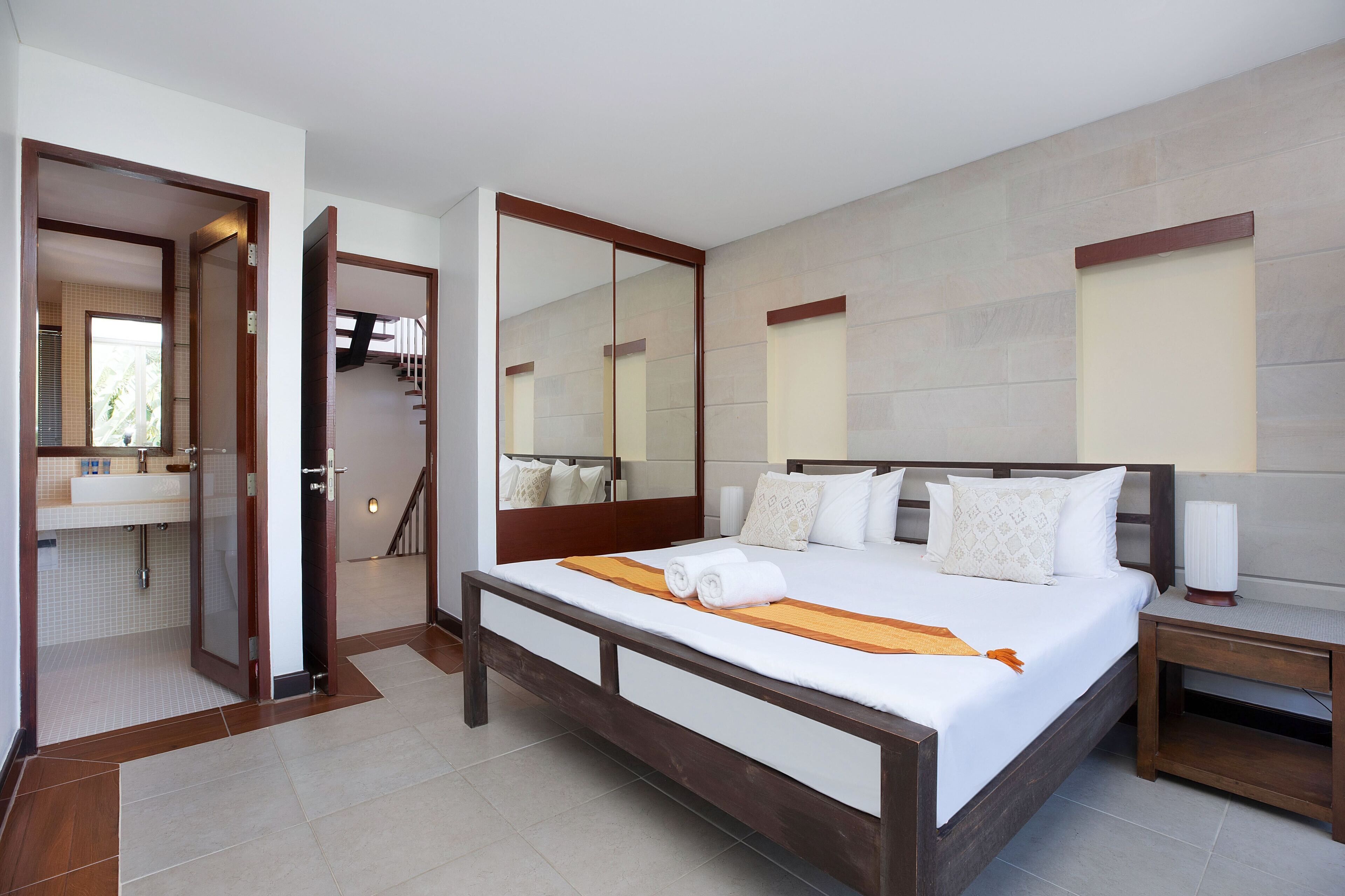 Una habitación de hotel con una cama grande, un cabecero de madera, una mesita de noche, un tocador, un espejo y un baño con lavamanos y ducha.