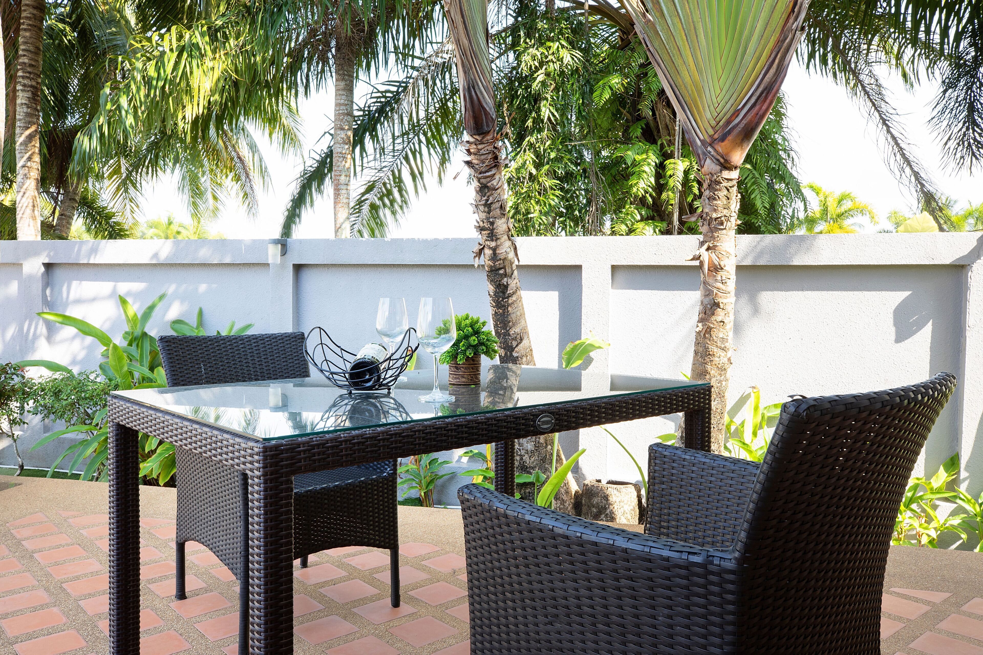 A glass-top table with wicker chairs, a hammock, and a white wall with greenery in the background.