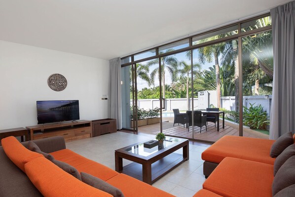 A modern living room with a flat-screen TV, orange sofas, a glass coffee table, and a view of palm trees through large sliding glass doors. - Private 3BR Pool Access Villa Argent 10 (Thep Krasattri)