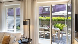 Ein modernes Wohnzimmer mit einer Couch, einem Beistelltisch mit Dekoration und einer großen Schiebeglastür, die auf eine Terrasse im Freien mit Esstisch und Stühlen führt.