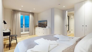 Ein Schlafzimmer mit einem großen Bett, einem Schreibtisch, einem Sessel, einer Kommode und Meerblick durch ein Fenster.