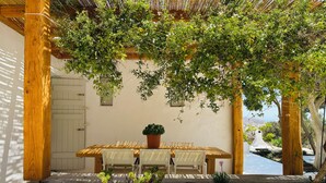 A covered outdoor dining area with a table and chairs, surrounded by greenery and a wooden pergola. - Stone Built Villa Ferretti #Unoblu (Mykonos)