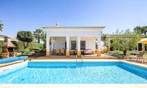 Un espace aménagé au bord de la piscine, doté d’un coin salon couvert, d’une piscine aux motifs géométriques réalisés avec des carreaux, et entouré de verdure.