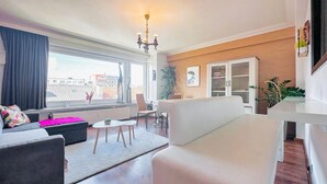 Ein modernes Wohnzimmer mit einer Couch, einem Esstisch und Stühlen. Es gibt ein großes Fenster mit Vorhängen, eine Kristallleuchter und ein Bücherregal.
